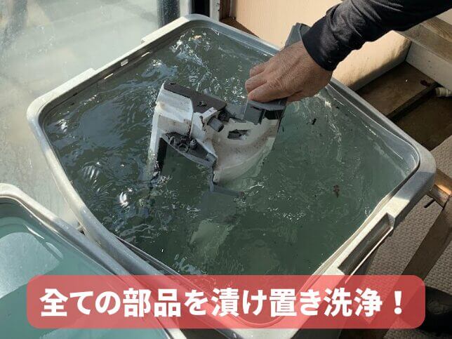 エアコン分解クリーニングで取り外した部品を水槽に浸け置き洗浄している様子。画面下には「全ての部品を漬け置き洗浄!」と表示。